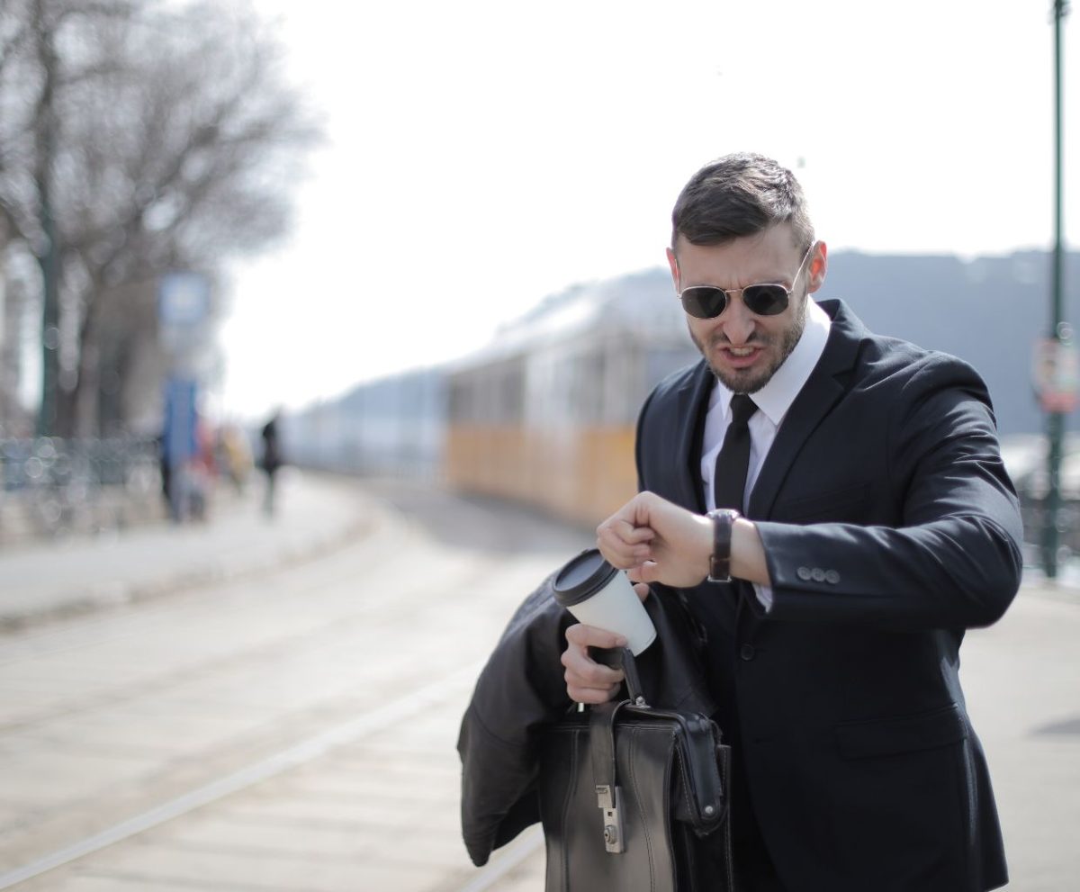 Hombre con traje y gafas de sol mirando su reloj mientras sostiene un maletín y una chaqueta