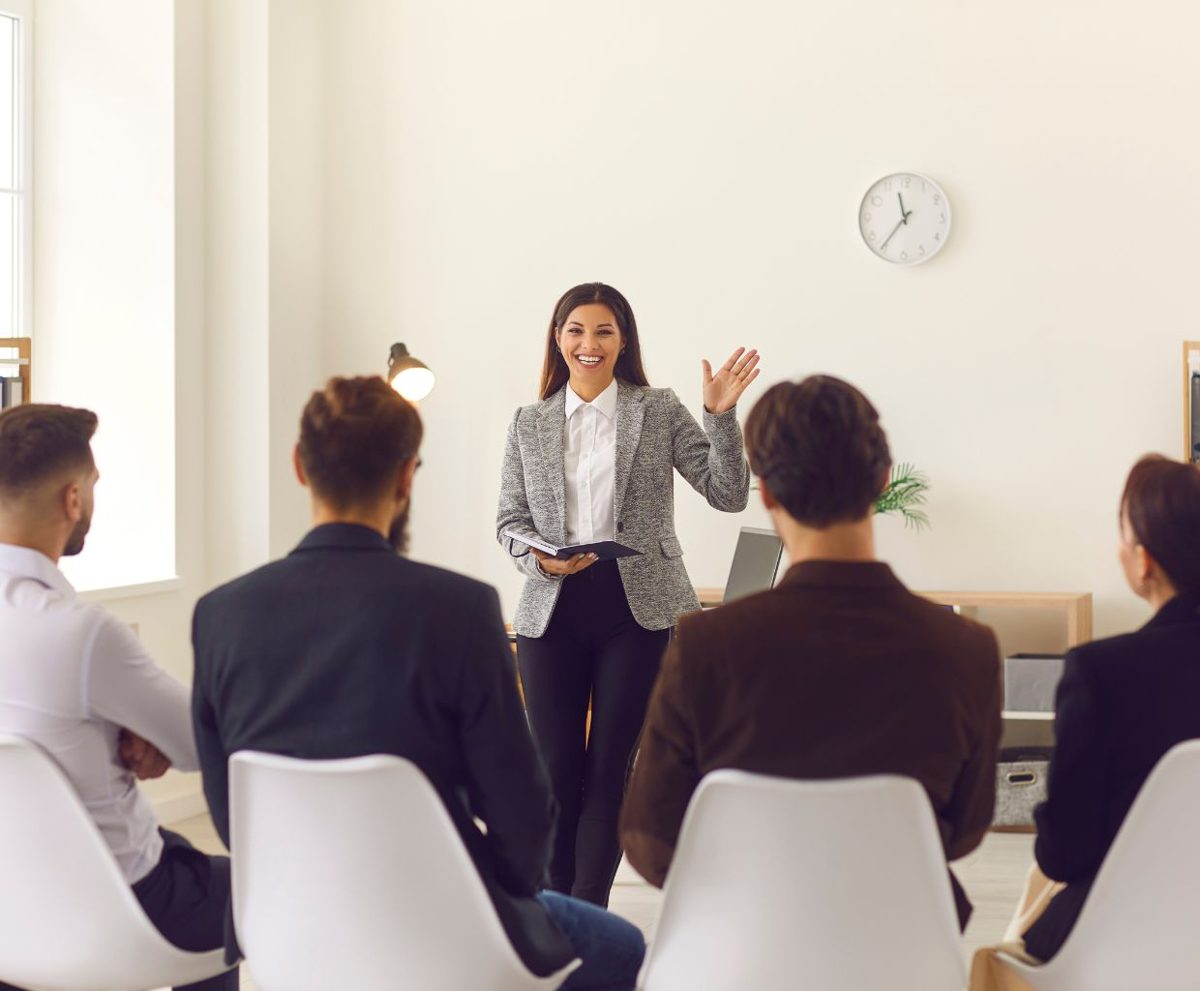 Profesora de pie explicando ante un grupo de personas sentadas en una sala
