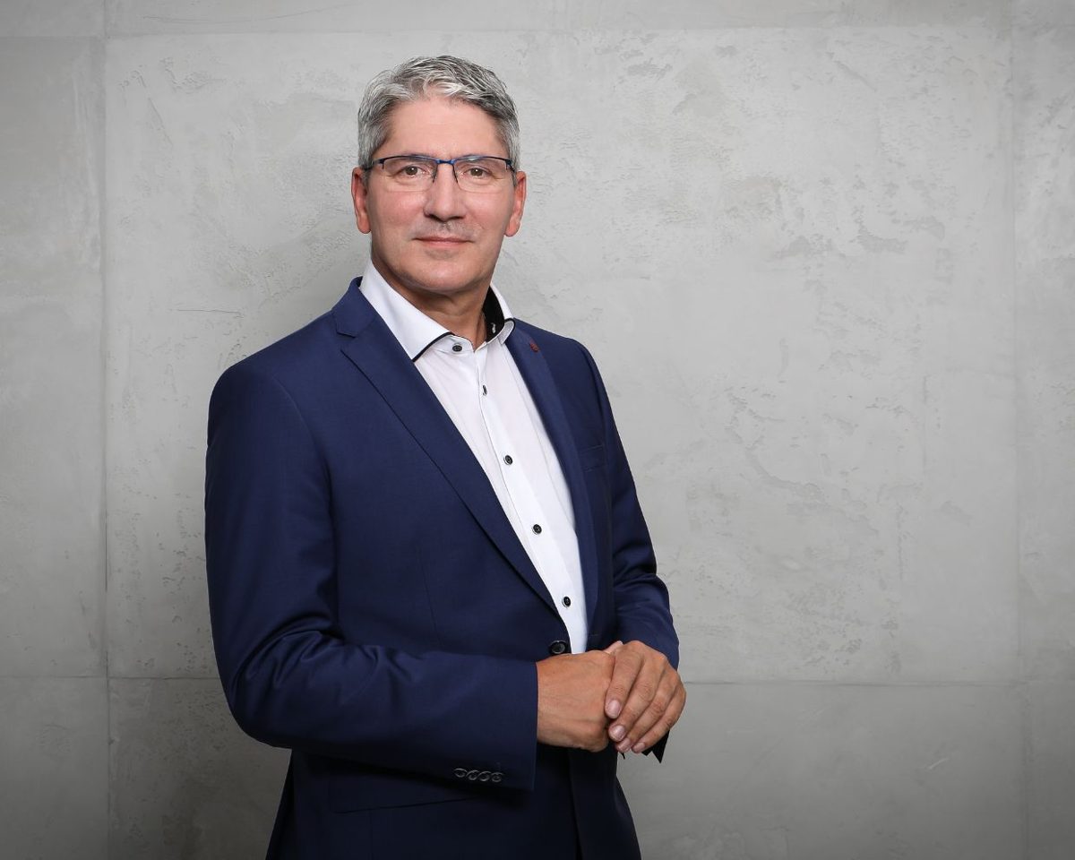 Hombre con traje azul y camisa blanca posando de pie con las manos juntas ante un fondo gris