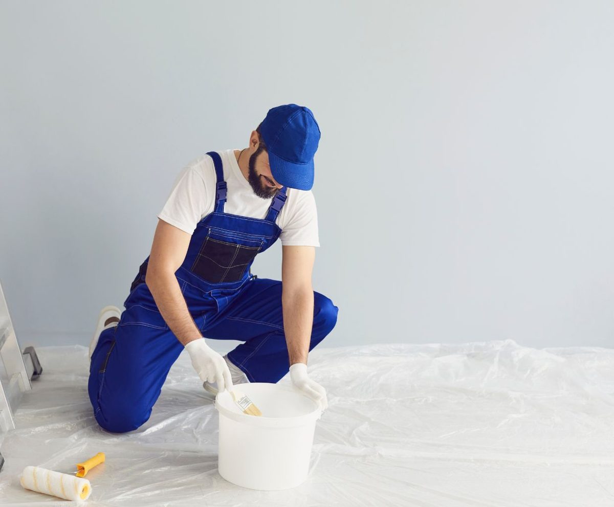 Trabajador con mono azul y gorra agachado junto a un cubo sobre un suelo cubierto con plástico