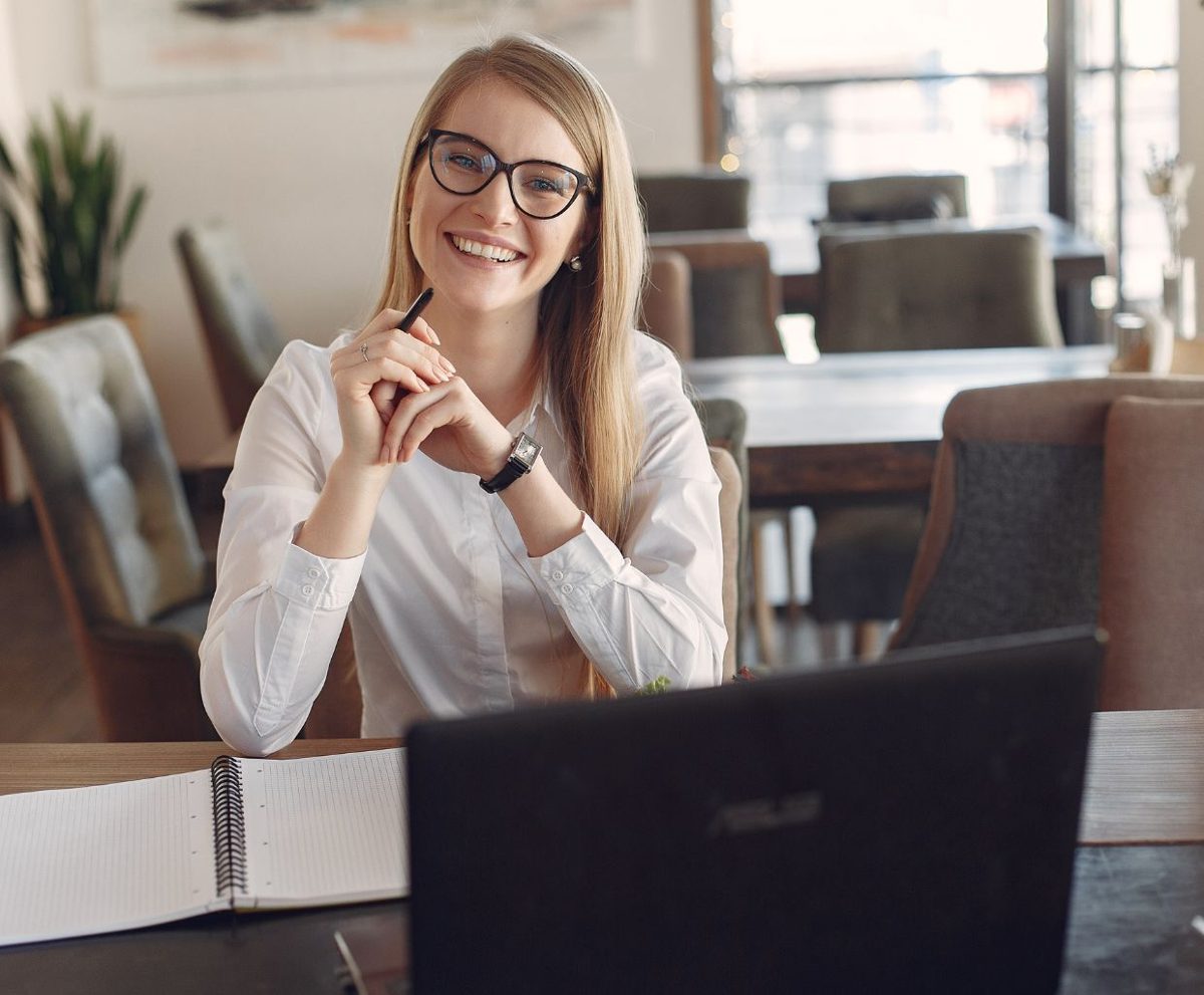 Mujer joven sonriente con gafas sentada ante su portátil en una mesa, lista para una videollamada