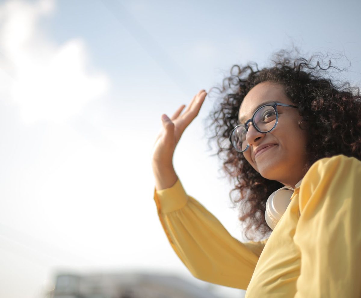Mujer de pelo rizado y gafas saludando con la mano ante un cielo azul