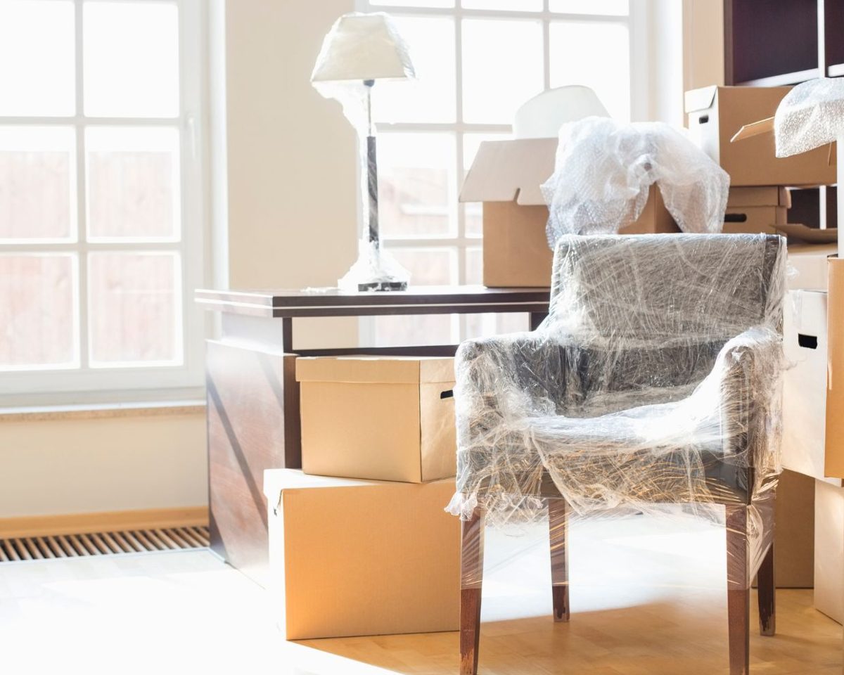Habitación con cajas de cartón de mudanza y una silla cubierta con tela junto a una ventana