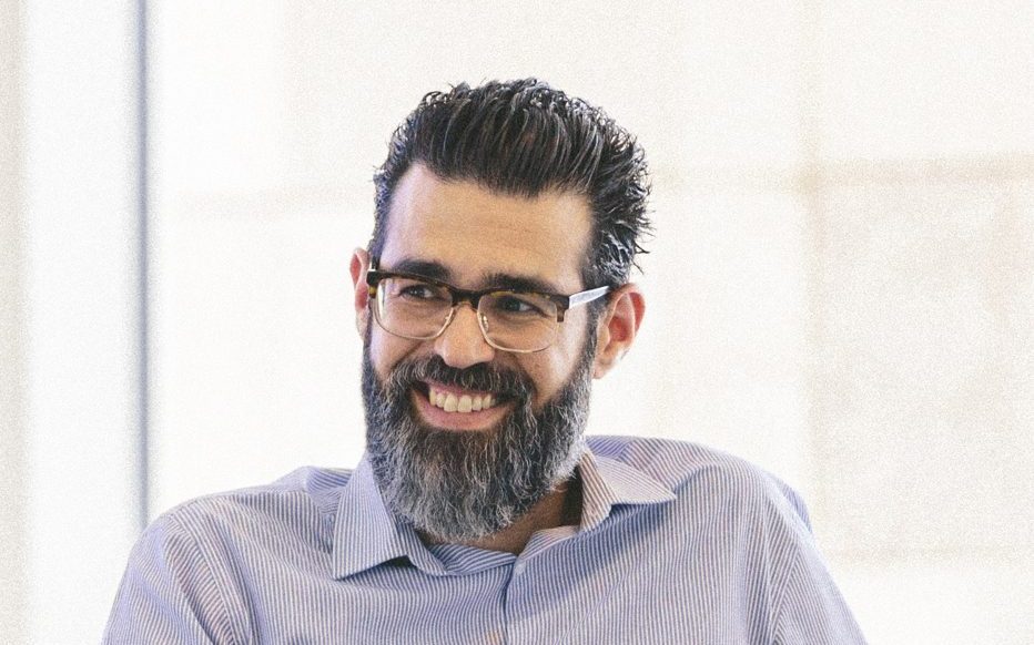 Hombre con barba y camisa azul sonriendo sentado en un sillón durante una conversación