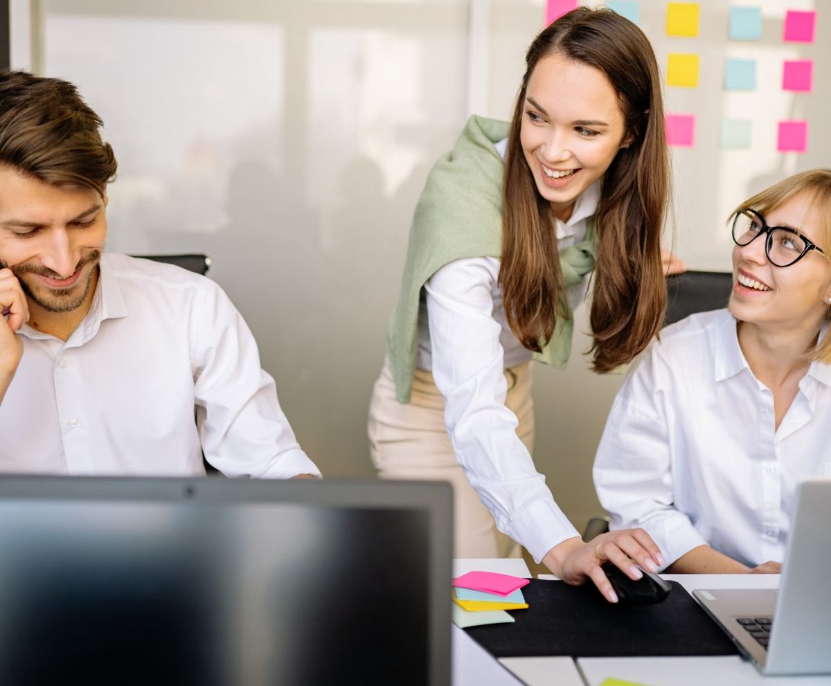 Equipo de compañeros sonriendo mientras una mujer les muestra algo en una pantalla en una oficina con notas adhesivas