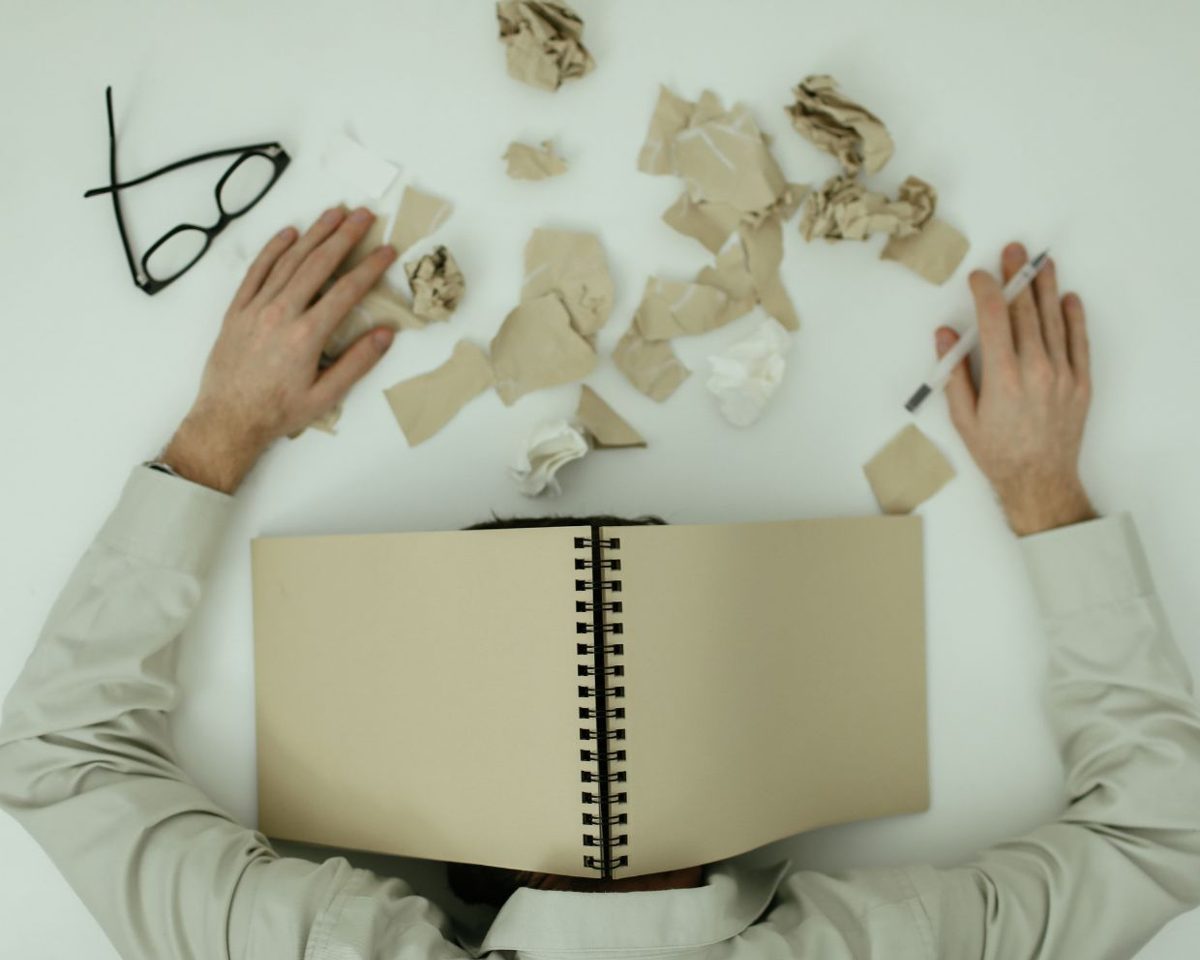 Persona sentada en un escritorio con las manos sobre la cara y un cuaderno cubriendo parte de la cabeza