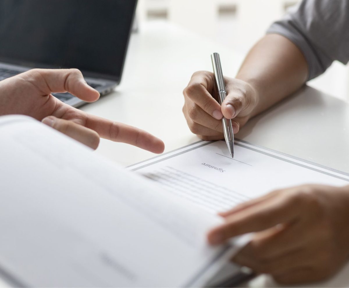 Persona firmando un documento mientras otra señala una línea con el dedo