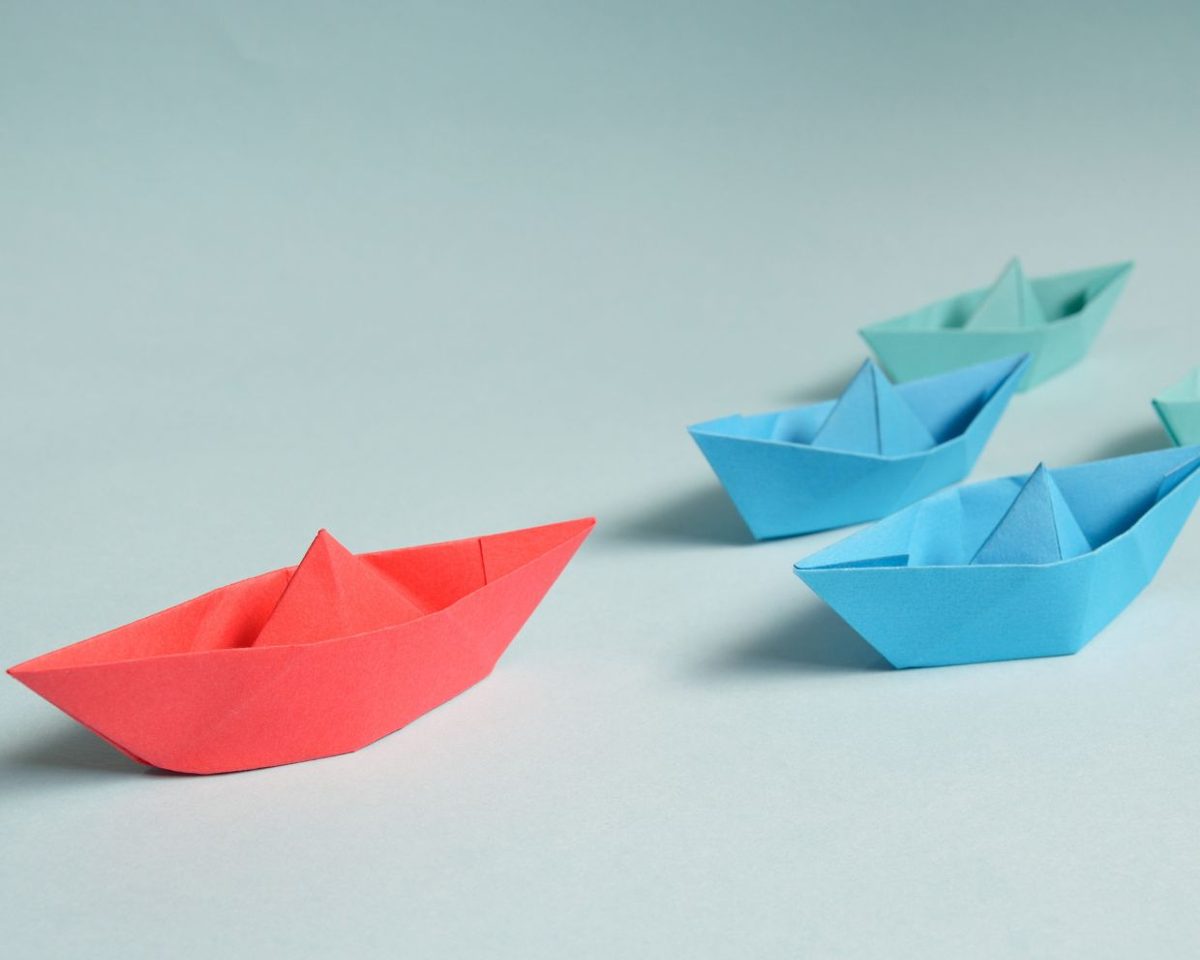 Barcos de papel de colores sobre una superficie azul, uno rojo mirando en distinta dirección