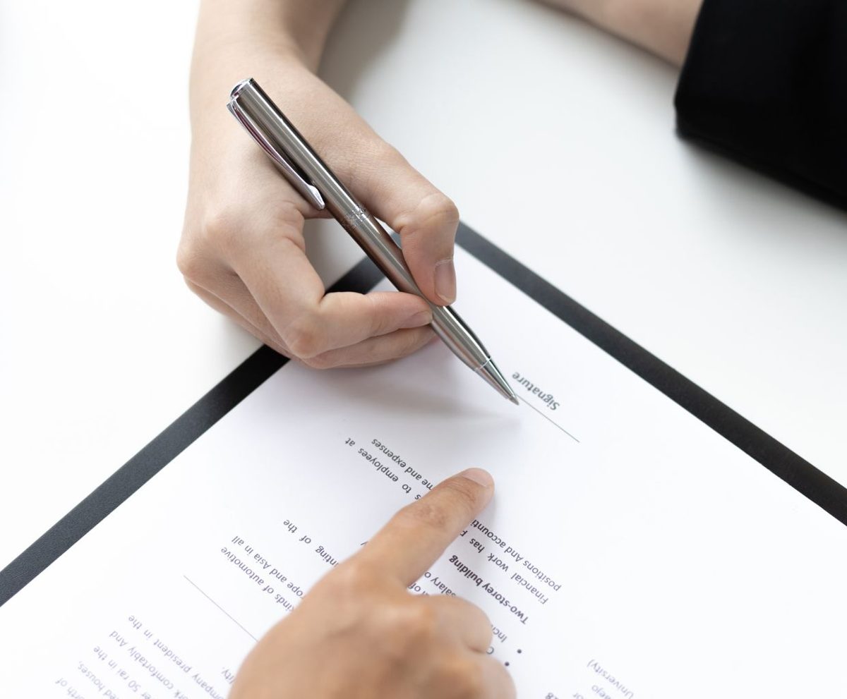 Mano con un bolígrafo firmando un documento sobre un portapapeles mientras otra mano señala la línea de firma