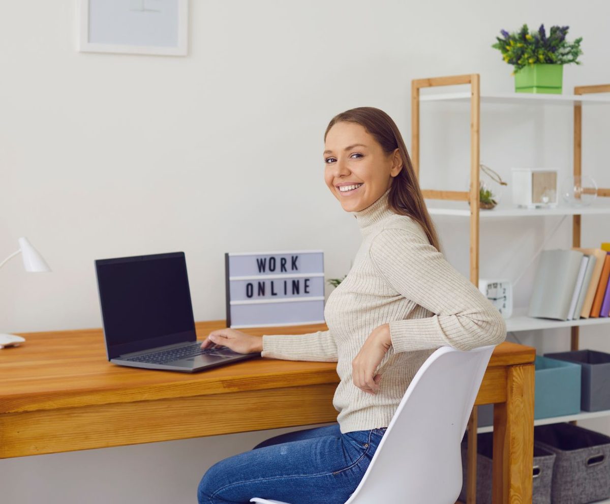 Mujer sonriente sentada en un escritorio de madera junto a un portátil con un cartel que dice Work Online